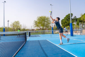 Full length photo of a young caucasian man jumping while playing pickleball in an outdoor facility